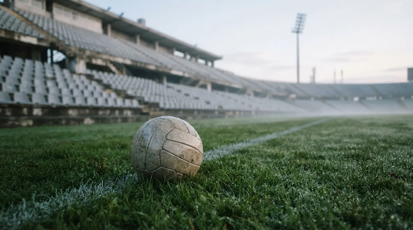 Ein Fußball liegt auf dem Rasen vor einer leeren Tribüne am frühen Morgen, Tau glänzt auf dem Gras.