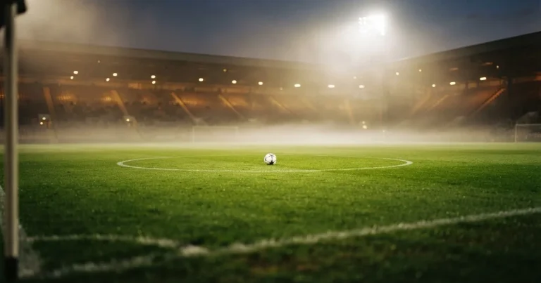 Ein Stadion unter Flutlicht aus der Ferne am Abend, hell erleuchtet, Nebel liegt über dem Rasen.