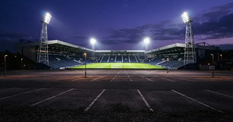 Ein Fußballstadion unter Flutlicht aus großer Entfernung gesehen, die Lichtkegel stehen im dunklen Abendhimmel über leeren Parkplätzen.